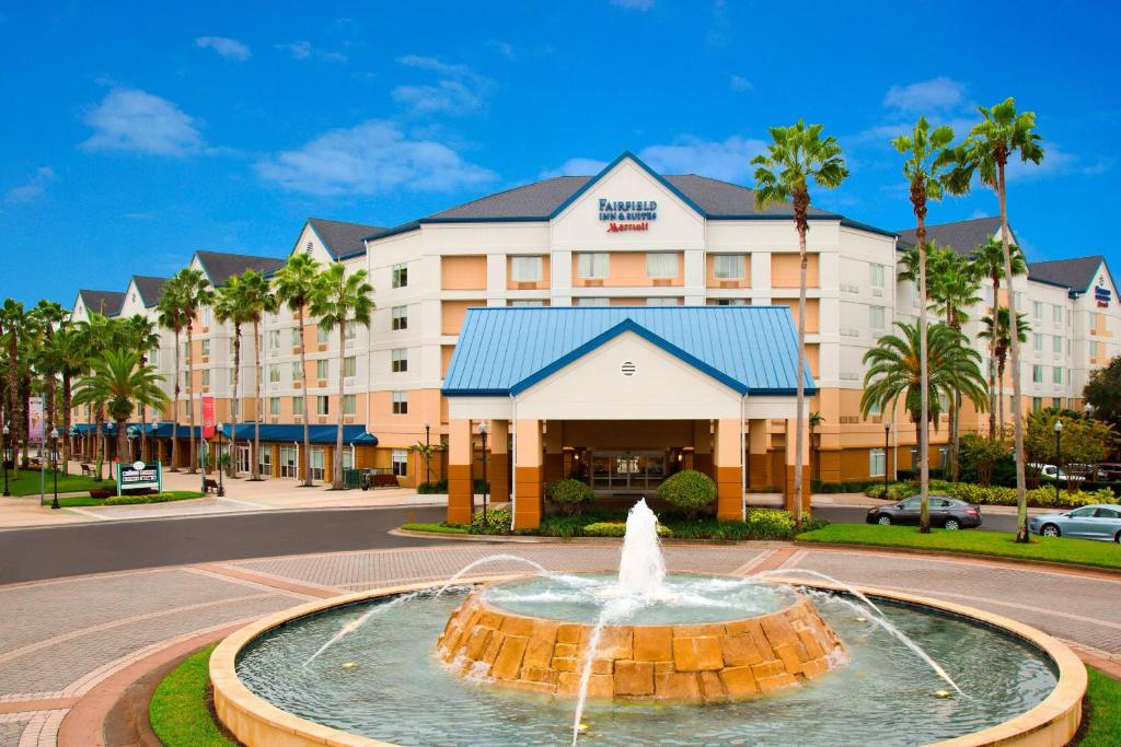 A multi-story Fairfield Inn & Suites by Marriott hotel with a blue roof entrance and a circular fountain in front, surrounded by palm trees and parked cars, under a clear blue sky.