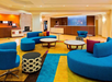 Modern hotel lobby with curved blue sofas, colorful area rugs, round tables, patterned ottomans, and a reception desk in the background.