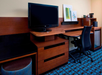 Hotel room workspace with a flat-screen TV, wooden desk, office chair, mini fridge, and framed artwork on a wall above patterned carpet.