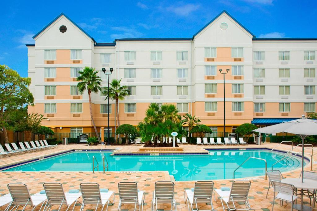 Outdoor swimming pool with lounge chairs, umbrellas, and palm trees in front of a multi-story hotel building under a clear blue sky.