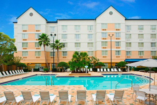 Outdoor swimming pool with lounge chairs, umbrellas, and palm trees in front of a multi-story hotel building under a clear blue sky.