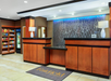 Hotel reception area with wooden front desk, abstract wall art, and two lamps. Snack shop on the left with shelves of packaged goods and a refrigerator.