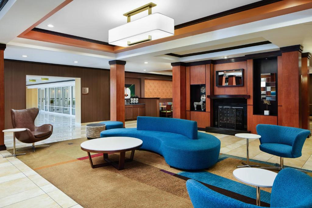 Modern hotel lobby with blue seating, a unique curved sofa, round tables, wood accents, and a fireplace. Bright lighting and a hallway visible in the background.