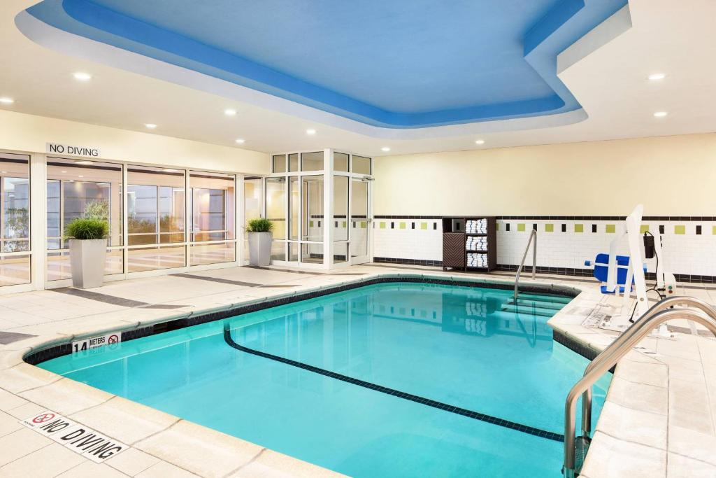 Indoor swimming pool with blue ceiling, chairs, and clean tiled floor. Towels are stacked near the entrance. No diving signs are visible.