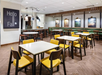 A modern cafe interior with tables, yellow chairs, and wall art. A sign reads "Hello Fall" on one wall.