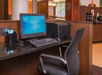 Hotel business center with a computer, printer, and office chair on a wooden desk. Nearby are coffee dispensers and stir sticks.
