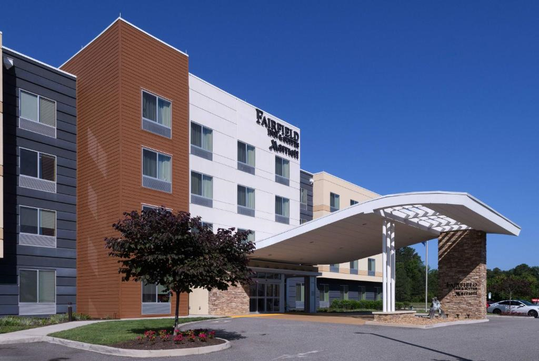 Modern hotel exterior with a covered entrance and a landscaped area featuring a small tree and flowers. The building has multiple floors with rectangular windows.