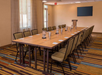 A conference room with a long table surrounded by chairs, notepads, pens, and water pitchers. A podium and flat-screen TV are visible in the background. Striped carpet covers the floor.