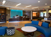 Hotel lobby with modern decor, featuring a blue sofa, round white table, and check-in desks in the background. Soft lighting and a small snack area on the left.