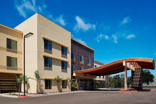 Modern hotel exterior with a three-story building, large windows, and an arched wooden entrance. Surrounding palm trees and a clear blue sky are visible.