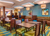 Hotel dining area with wooden tables and chairs, colorful carpet, blue walls, and framed art. Ceiling lights illuminate the space.
