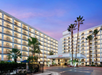 A large hotel building with balconies is illuminated at dusk, surrounded by palm trees. The entrance is bright and welcoming.