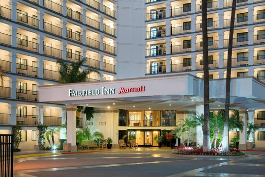 Exterior view of a multi-story Fairfield Inn by Marriott hotel, featuring balconies, illuminated windows, and a central entrance surrounded by palm trees.
