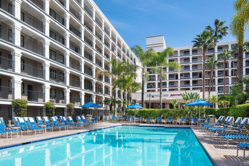 Outdoor hotel pool surrounded by lounge chairs and umbrellas, with a large hotel building and palm trees in the background.