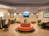 A hotel lobby with a round orange sofa, green chairs, tables, framed artwork, and a wall-mounted TV displaying a blue screen.