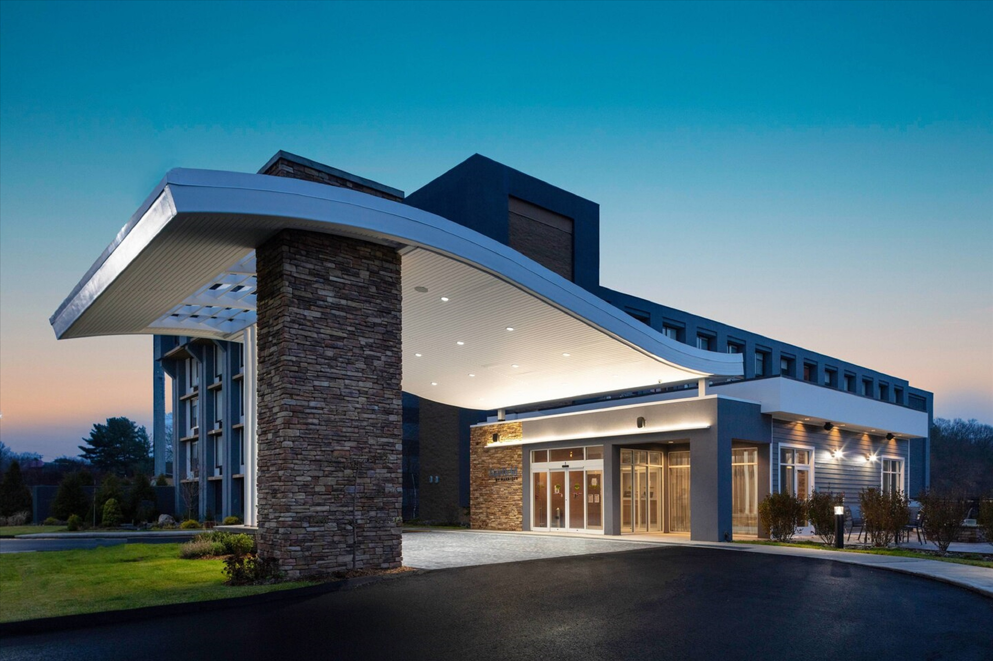 Modern hotel entrance with a curved overhang, stone pillars, and glass doors, set against a clear evening sky.
