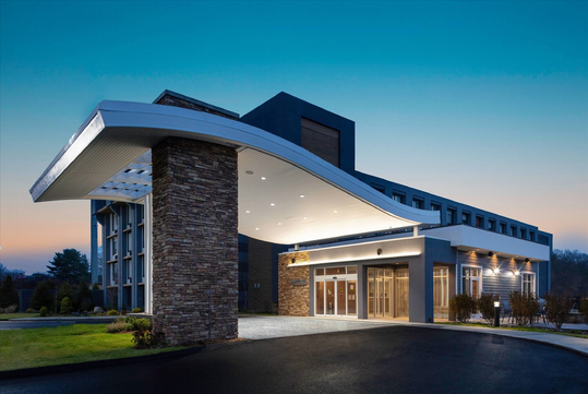 Modern hotel entrance with a curved overhang, stone pillars, and glass doors, set against a clear evening sky.