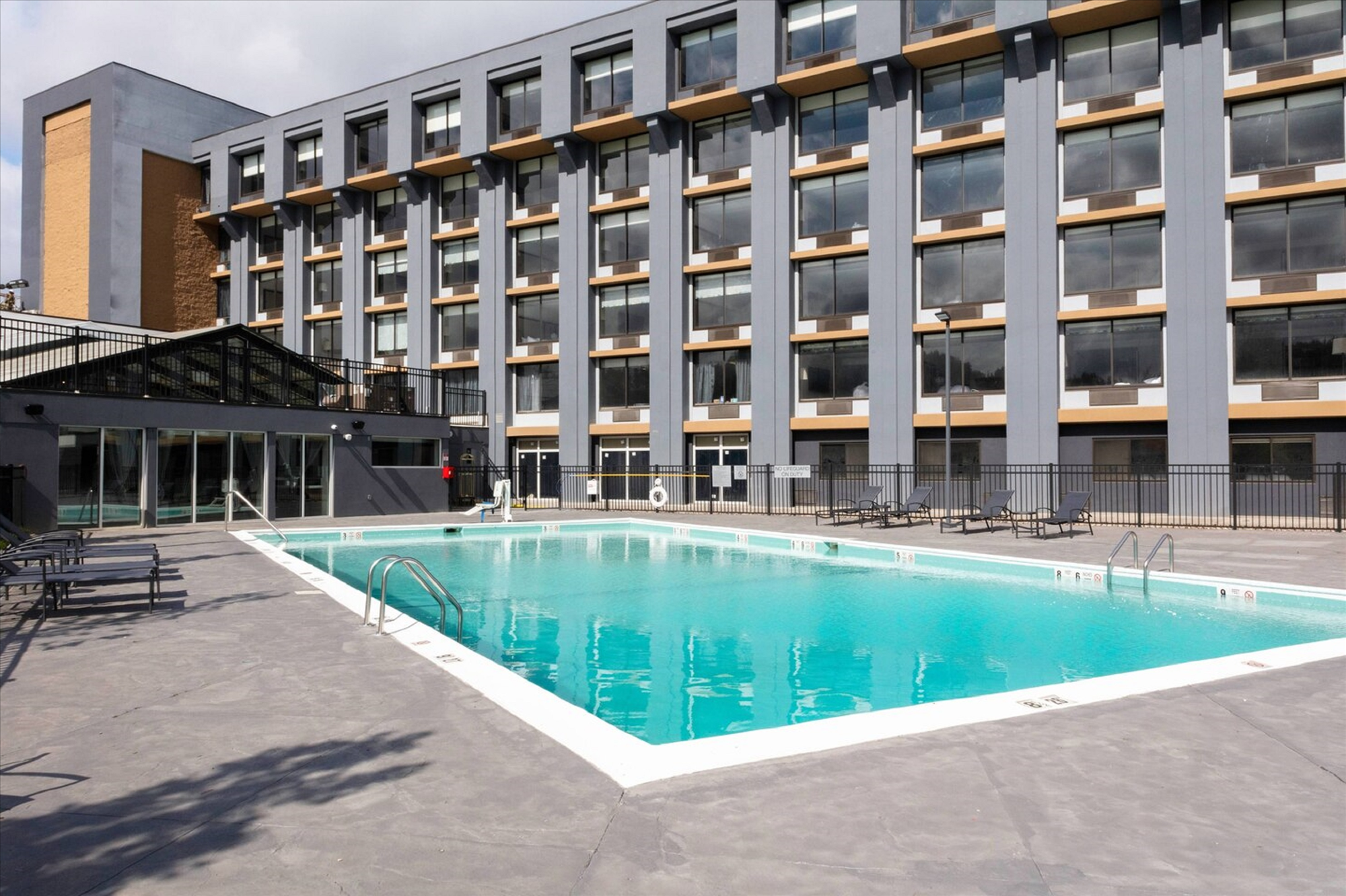 Outdoor swimming pool surrounded by a concrete deck in front of a multi-story building with numerous windows.
