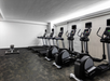 A gym with five elliptical machines, five treadmills, and a lone rowing machine on a dark carpeted floor, beneath a white ceiling and a bright overhead light.