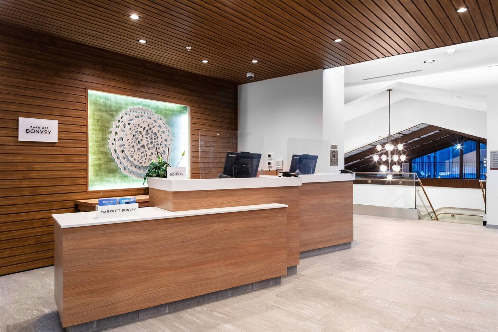 Hotel reception area with a wooden desk and wall, modern ceiling lights, and a staircase to the right. Two computer monitors are on the counter.