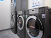 Row of industrial washing machines in a laundromat setting, with silver exteriors and digital displays.