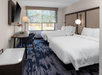 Hotel room with two double beds, a desk with a lamp and chair, mounted TV, and a large window with a nature view. Carpet has a blue pattern. Walls are neutral-colored.