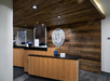 Hotel reception area with wooden counters and ceiling, featuring a circular metal wall art piece. Two computer monitors are on the counters, with acrylic barriers for separation.