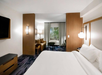 Hotel room with a large bed, two bedside lamps, a flat-screen TV, a sofa, a coffee table, and curtains revealing a window view. Walls and decor are neutral-toned.