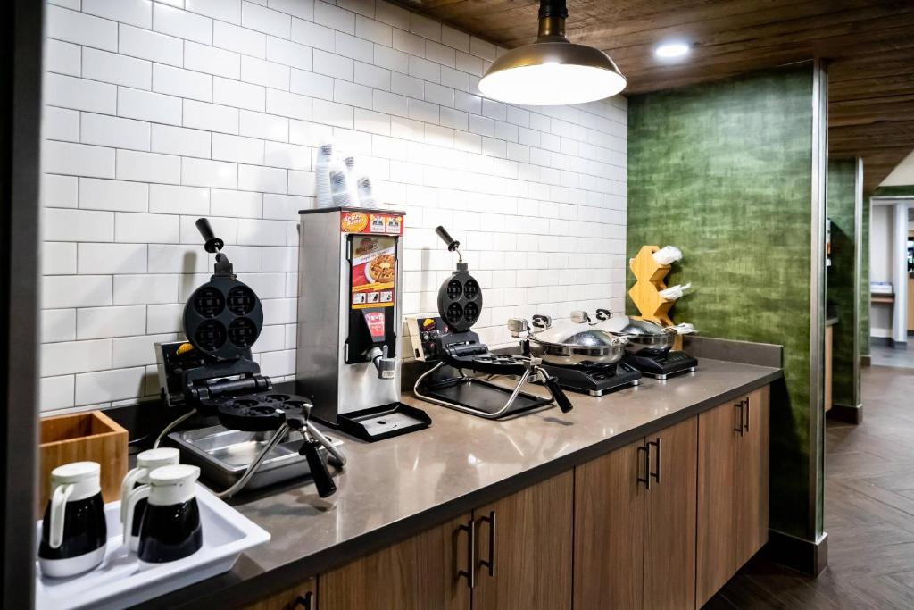 Hotel breakfast area with countertop waffle makers, waffle batter dispenser, condiment bottles, and stacked cups under a pendant light against a tiled wall.