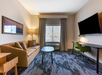 Hotel room with a couch, coffee table, wall-mounted TV, desk, and green chair. A large window with drawn curtains is in the background. Decor features neutral colors and modern design.