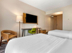Hotel room with two neatly made beds, a wall-mounted TV, a desk with a green chair, a brown armchair, and a floor lamp, featuring a modern and minimalist design.