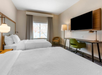 Hotel room with two beds, a large wall-mounted TV, a desk with a green chair, a floor lamp, and a window with curtains.