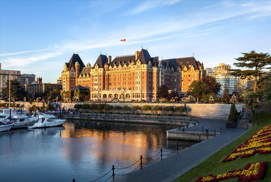 A large historic hotel with a Canadian flag, bordering a calm waterfront, surrounded by trees and a manicured garden, with several white boats docked nearby under a blue sky.