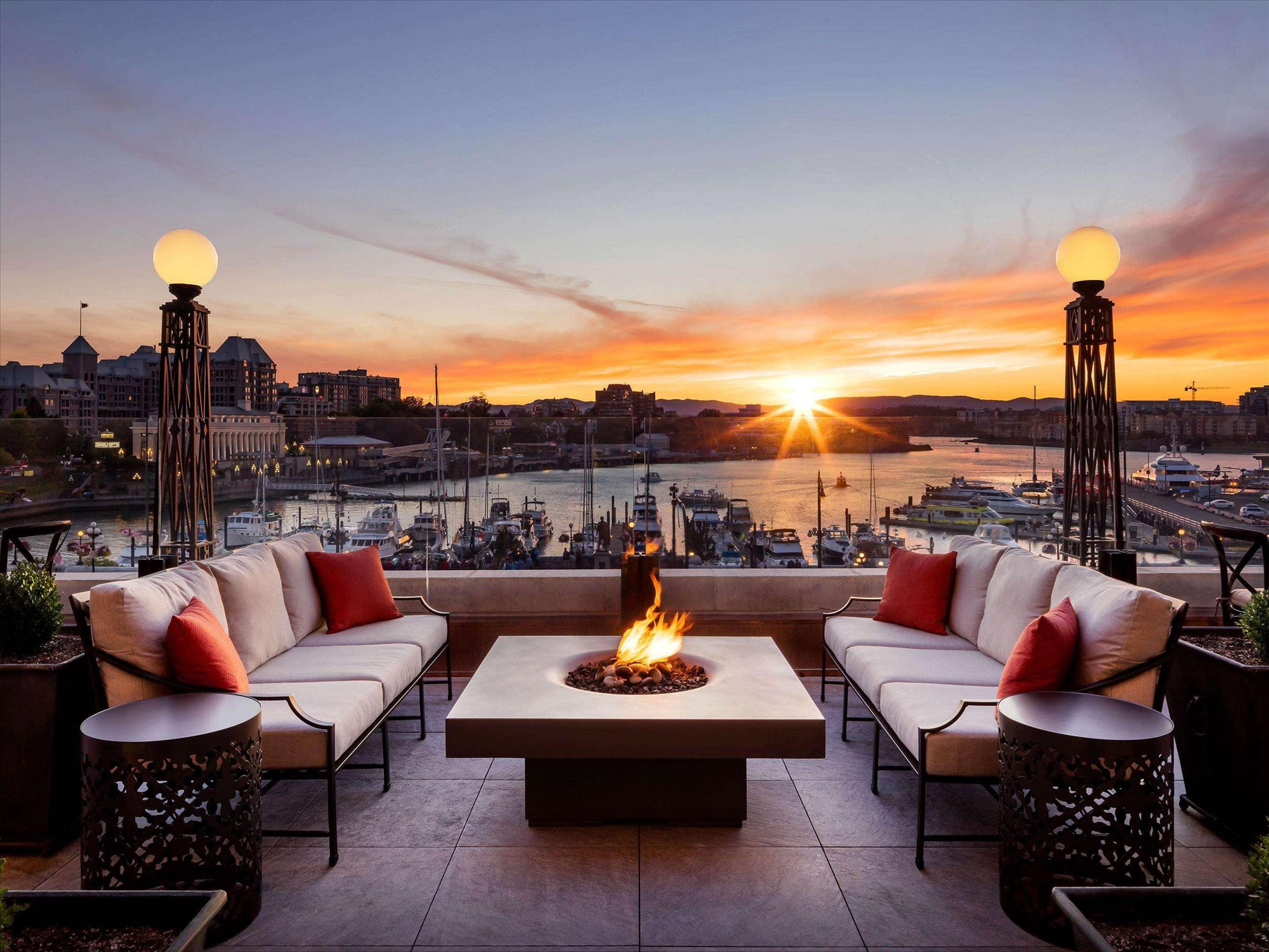 Outdoor patio with a square fire pit, surrounded by cushioned seating. The sunset view includes a harbor with boats and buildings in the background.