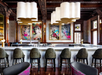 A stylish bar with a white countertop, modern pendant lights, and green bar stools. Colorful artwork depicting women adorns the wooden wall behind the counter.
