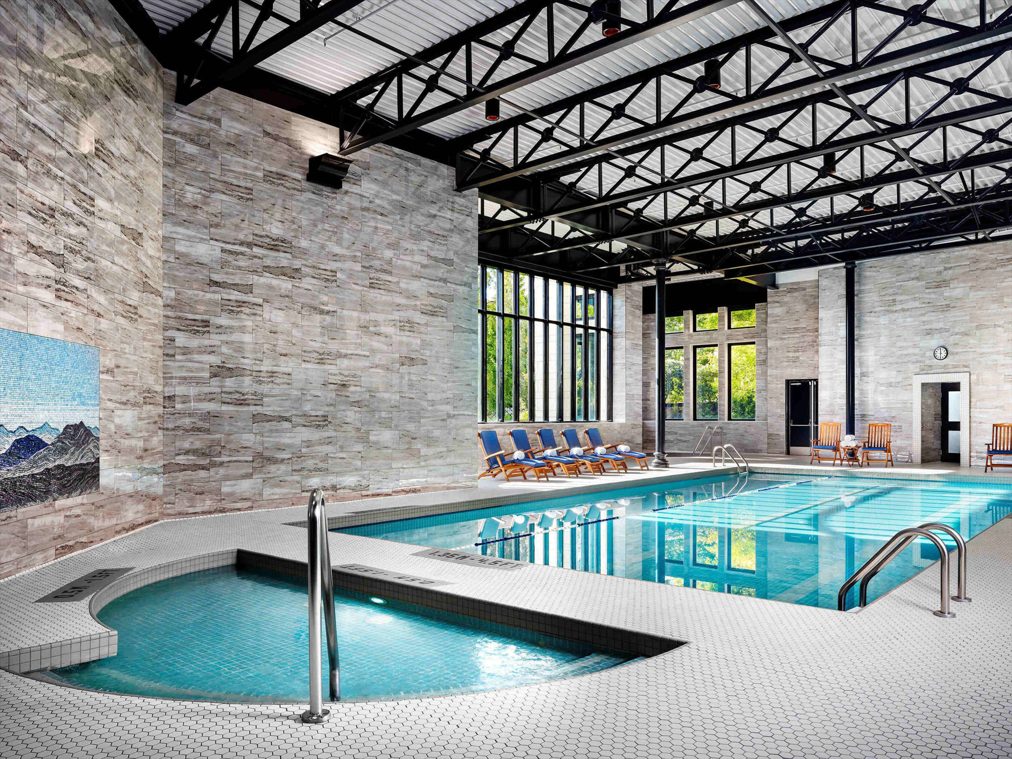 Indoor swimming pool area with a large pool and a smaller round pool. Several lounge chairs are arranged along the far wall near large windows allowing natural light inside.