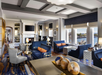 Elegant hotel lounge with blue sofas, armchairs, and a fireplace. Large windows overlook a waterfront view. Decorative accents include mirrors and ornamental spheres on a table.