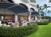 Outdoor restaurant with white tablecloths, wooden chairs, and blue awnings. Surrounded by greenery and palm trees, adjacent to a landscaped lawn.