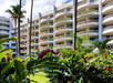 A multi-story hotel with curved balconies adorned with flowers and plants, surrounded by tall palm trees and lush greenery.