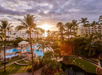 Aerial view of a tropical resort at sunset, featuring palm trees, a pool, surrounding greenery, and ocean in the background.