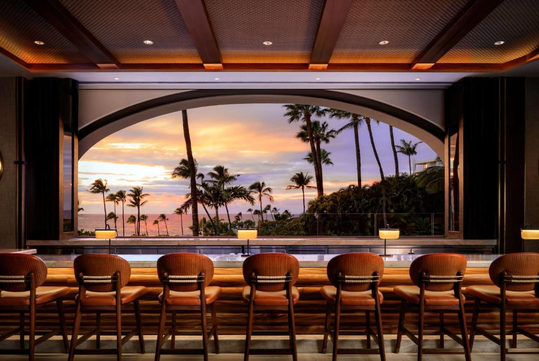 View of a tropical sunset over the ocean framed by an arched window, with a row of empty bar stools in the foreground.