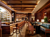 A stylish indoor bar area with a long counter, high wooden stools, and cozy seating. Warm lighting and intricate woodwork create an inviting atmosphere. Large windows reveal a view of a building outside.