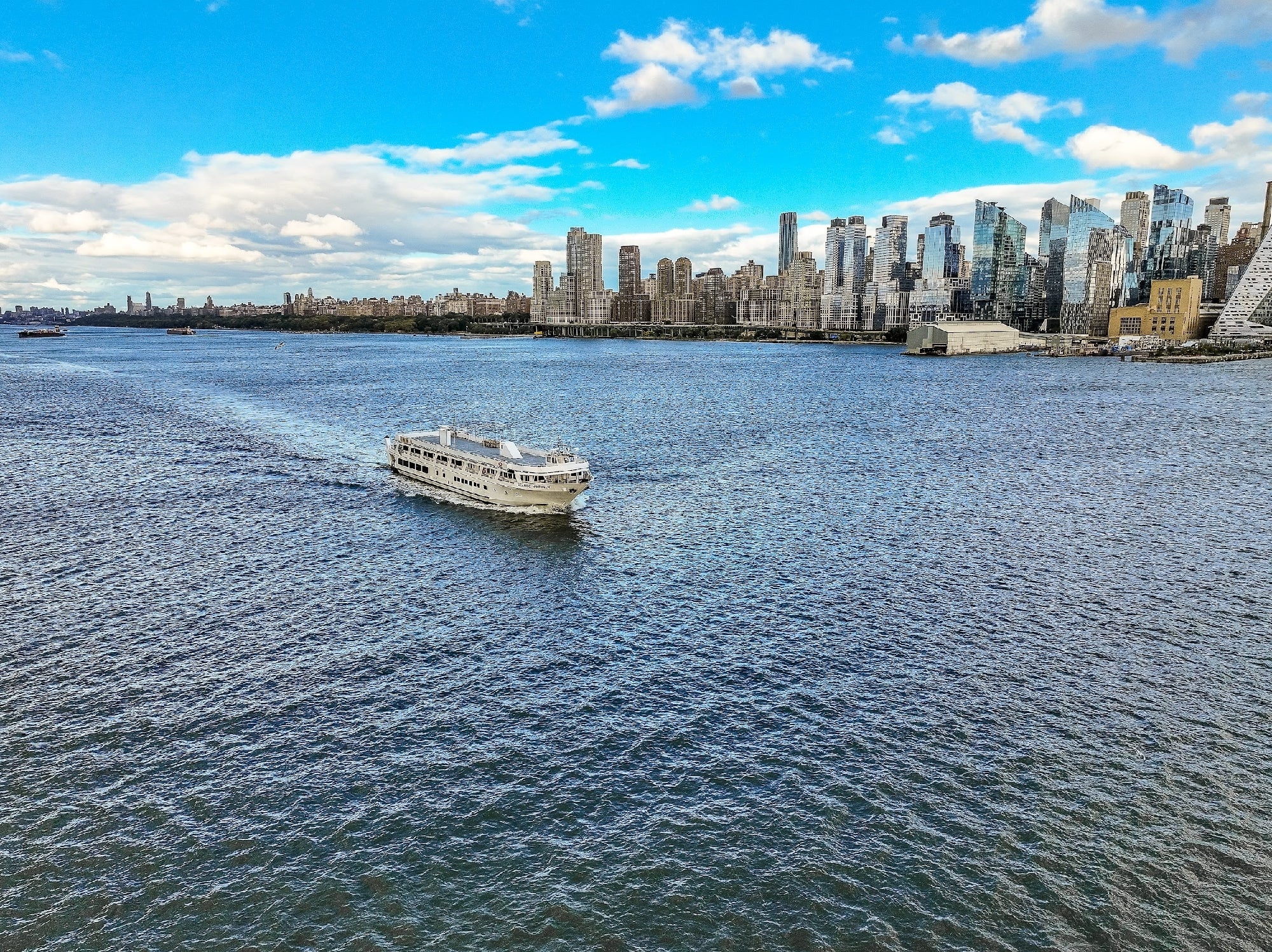 A white ferry boat travels across a wide river with a city skyline of modern high-rise buildings in the background under a partly cloudy sky.