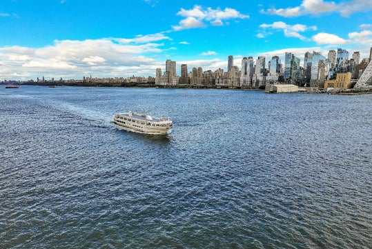 A white ferry boat travels across a wide river with a city skyline of modern high-rise buildings in the background under a partly cloudy sky.