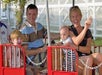 A family of four sits on a red amusement park train ride, with the parents smiling and the children sitting on their laps.