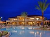 Night view of a resort with a large illuminated pool, surrounded by lounge chairs and umbrellas, with a two-story building and palm trees in the background.
