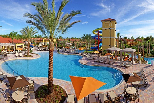 A large outdoor pool with sun loungers and umbrellas, surrounded by palm trees. A colorful water slide tower is in the background under a clear blue sky.