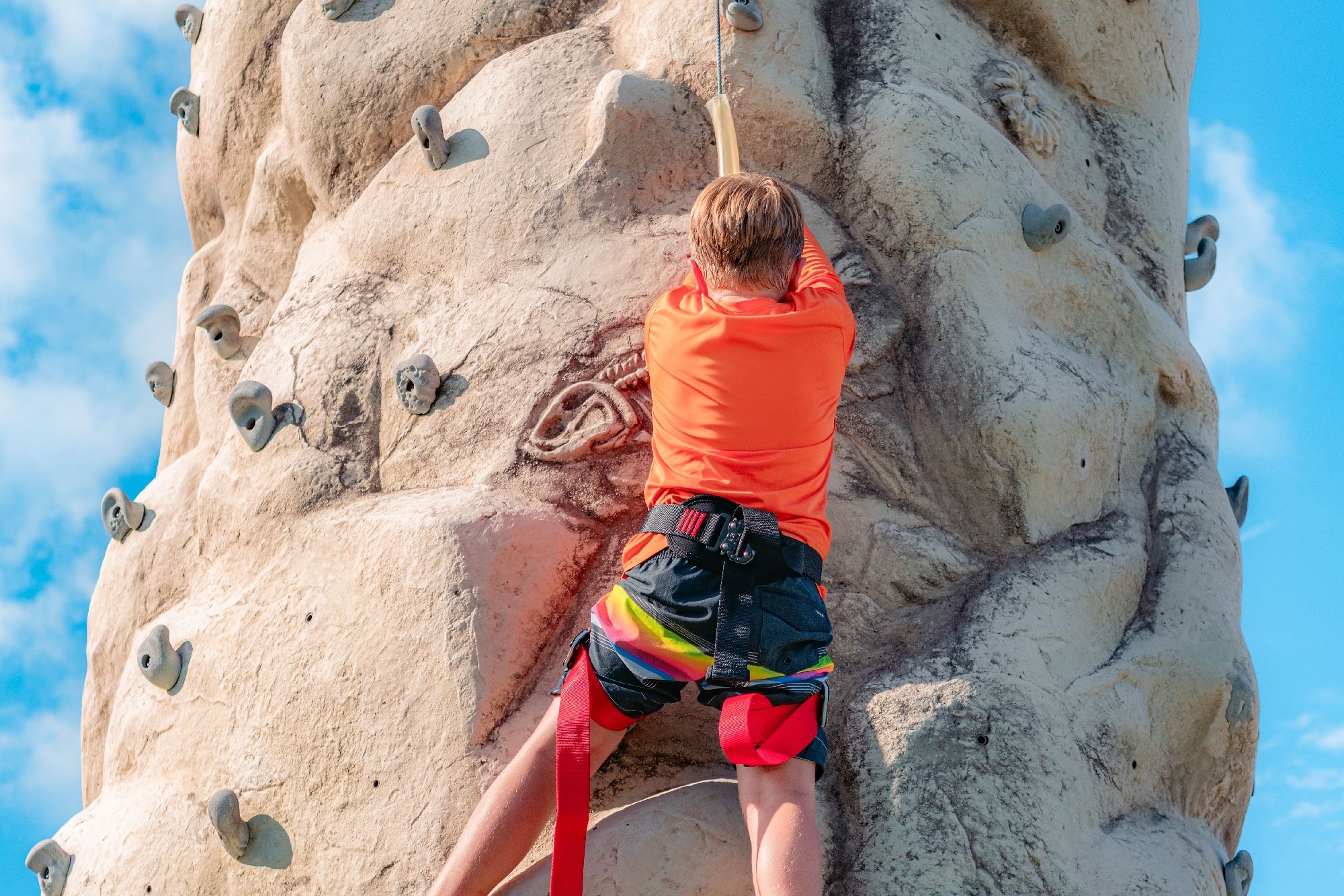 A young boy wearing a harness climbs an outdoor artificial rock wall, reaching for a hold under a blue sky.