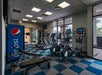 A gym room with treadmills, exercise bikes, a weight bench, and a Pepsi vending machine, featuring large windows and blue-and-gray checkered flooring.