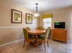 A small dining area with a round wooden table, four green cushioned chairs, a TV on a wooden cabinet, two leaf-themed wall art pieces, and a window with a green valance.